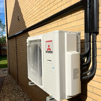 A Mitsubishi Heavy Industries inverter outdoor unit on wall brackets, with several pipe runs in black trunking rising up the brickwork to the eaves