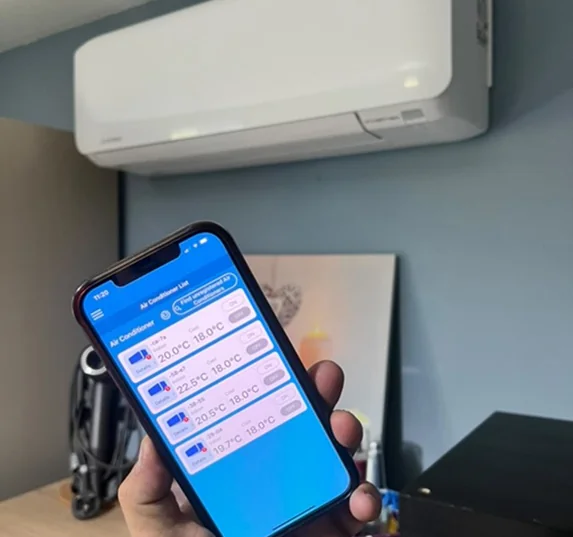 A phone showing an air conditioning control app with four rooms listed, held up in front of a wall-mounted indoor unit